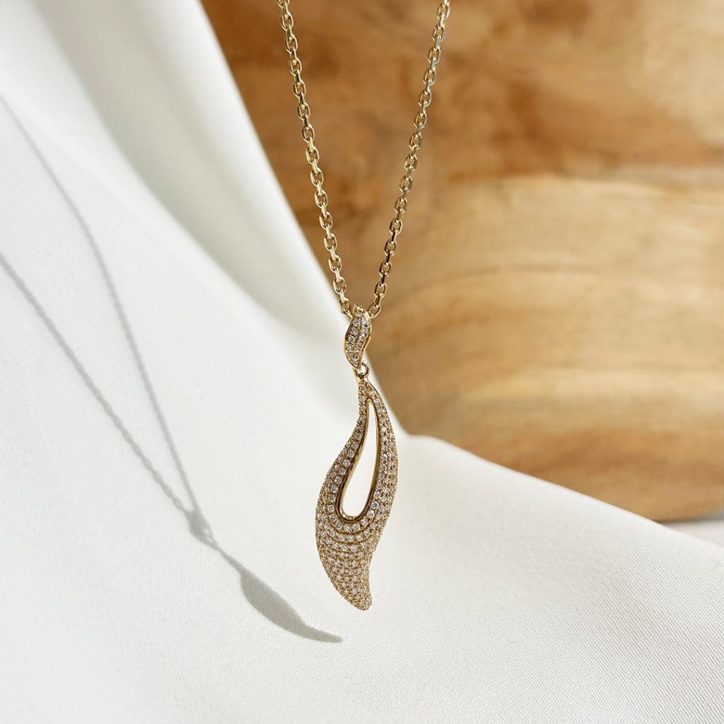 Eleganter, geschwungener Anhänger aus Gelbgold, vollständig mit funkelnden Diamanten besetzt. Getragen an einer Gelbgold-Kette, stilvoll fotografiert vor einer Holzwand.