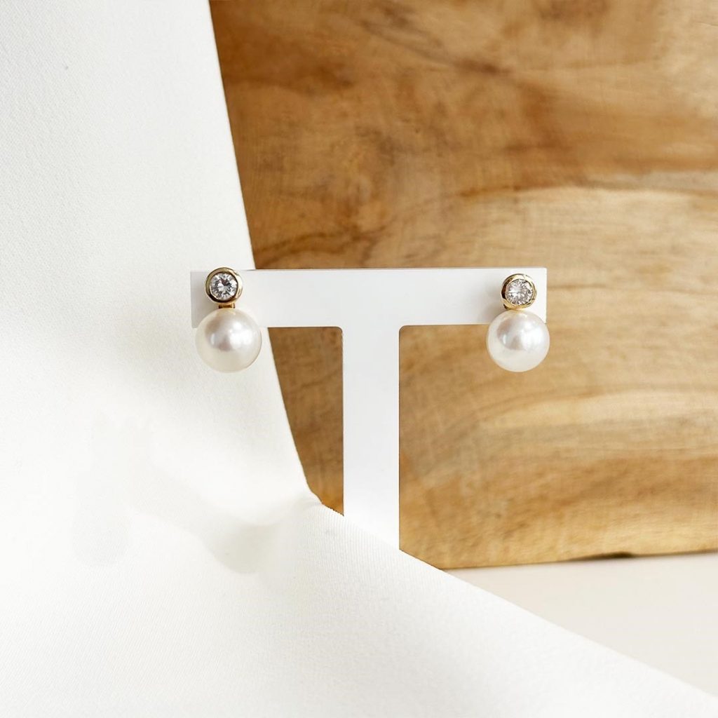 Ohrstecker mit Süßwasserperle und Brillant in 585 Gelbgold, präsentiert auf einem Schmuckaufsteller vor einer eleganten, hellen Holzwand. Artikelnummer 15989